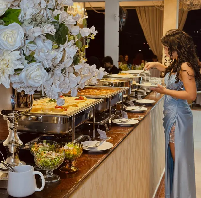 Convidada de vestido azul se servindo no buffet de jantar, entre rechauds, saladas em taças e flores brancas