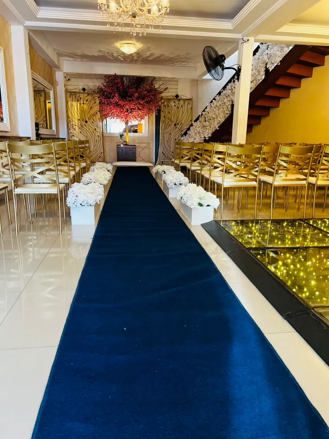 Corredor da cerimônia com tapete azul, cadeiras douradas, vasos de flores brancas e árvore de flores vermelhas no altar