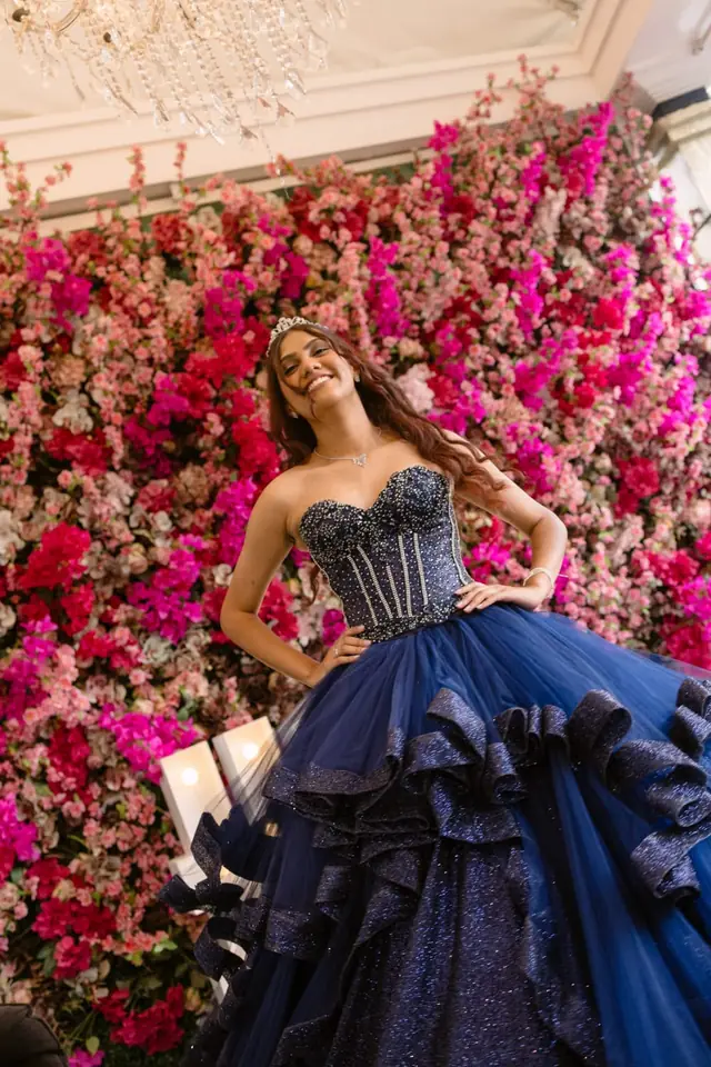 Debutante de vestido azul-marinho com corpete de brilhos e tiara, diante do painel de flores rosa e pink sob o lustre de cristal