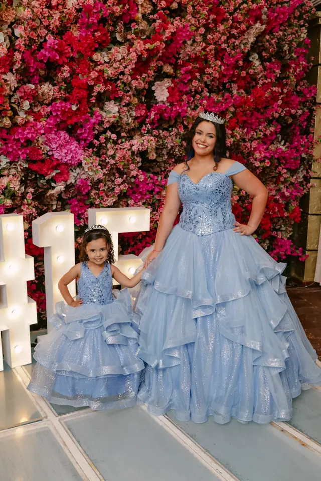 Jovem e menina de vestidos azul claro e tiara, diante do painel de flores e do número 15 luminoso