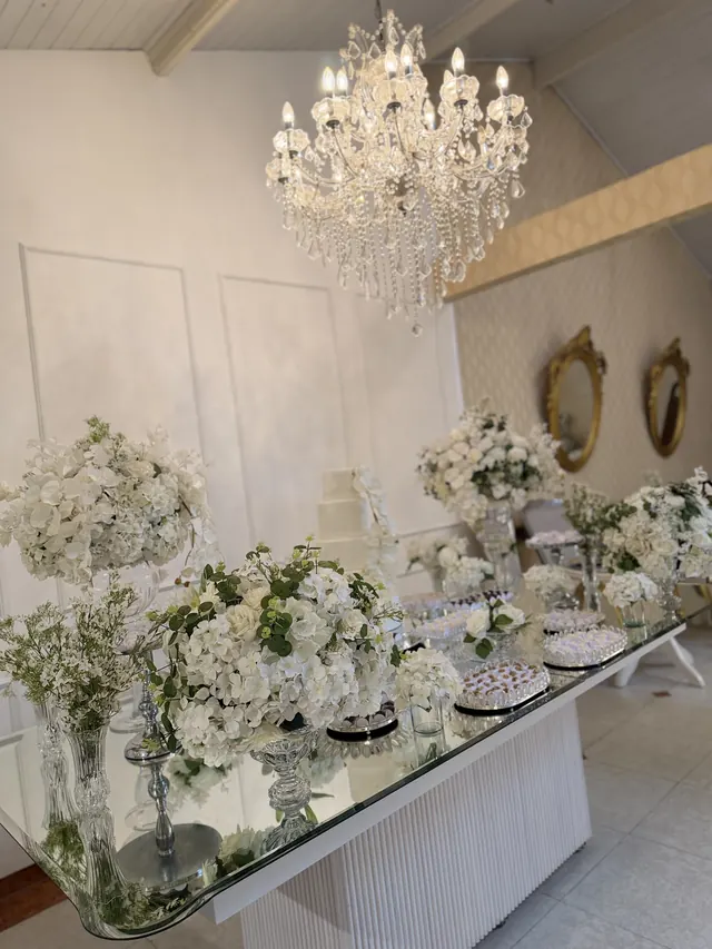 Mesa de doces com flores brancas, bolo e espelhos, sob o lustre de cristal do salão do Buffet Melvalen