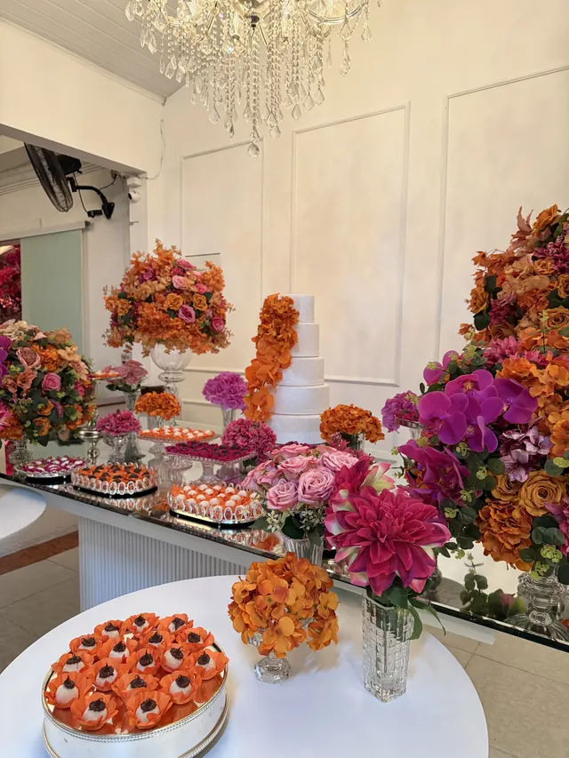 Mesa de doces com flores laranja e rosa e bolo de vários andares ao fundo
