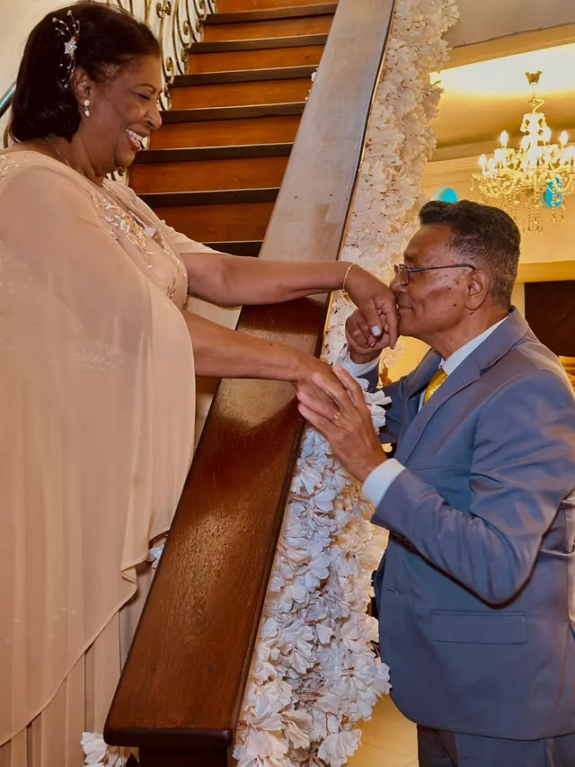 Casal em uma escada de madeira com o corrimão decorado com flores brancas e um lustre de cristal ao fundo