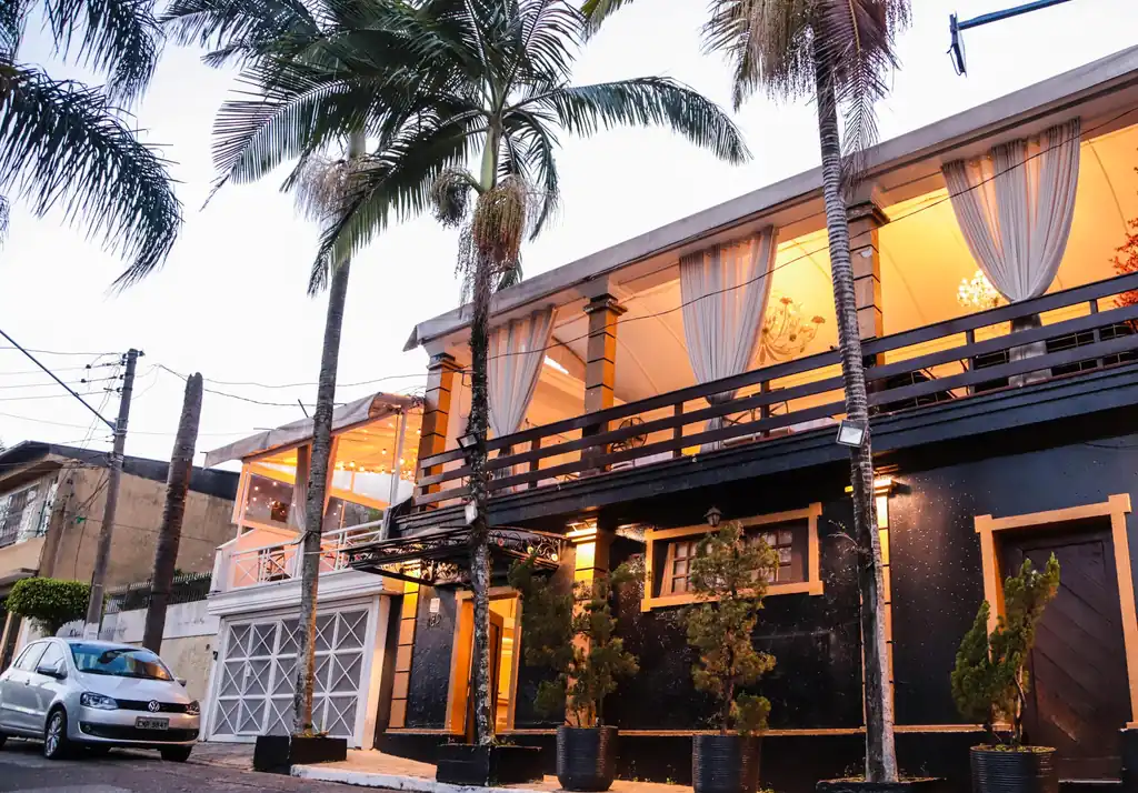 Fachada do Buffet Melvalen ao entardecer, sobrado escuro com varanda iluminada e palmeiras