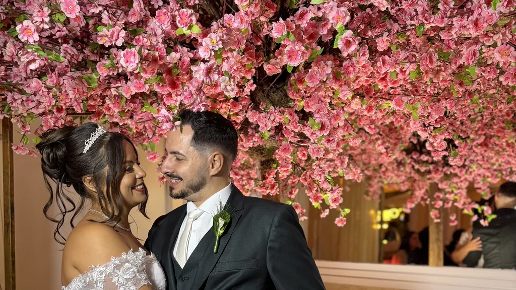 Casal de noivos, ela de vestido branco e tiara, ele de terno escuro, se olhando sob uma árvore de flores rosa no Buffet Melvalen