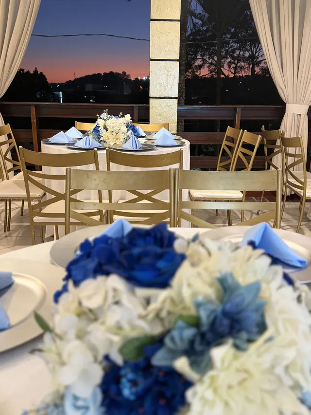 Mesa posta com arranjo de flores azuis e brancas e cadeiras douradas, com o céu do entardecer ao fundo