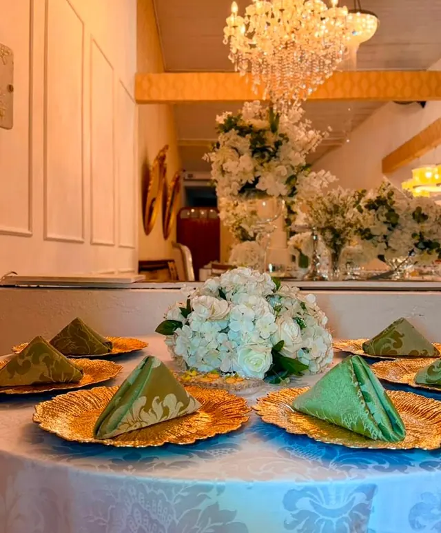 Mesa posta com sousplat dourado, guardanapos verdes dobrados e arranjo de flores brancas sob lustre de cristal