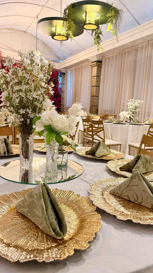 Mesa posta com sousplat dourado, guardanapo verde-oliva e arranjo de flores brancas no salão