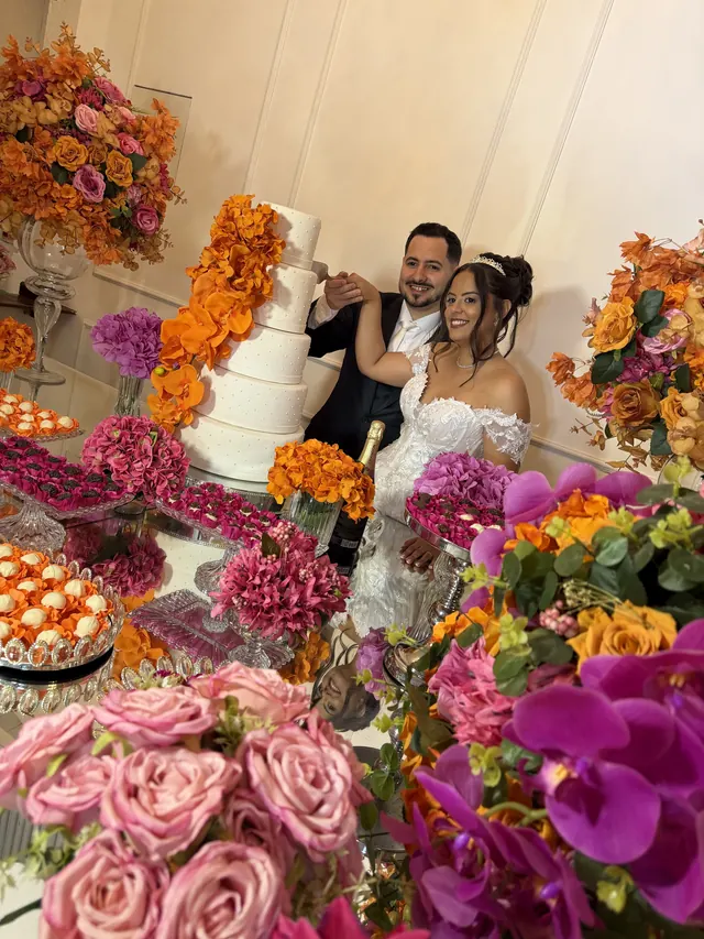 Noivos sentados ao lado de um bolo de vários andares, cercados por mesas de doces com flores laranja e rosa