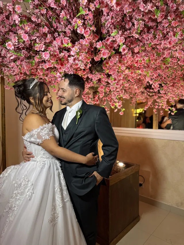 Casal de noivos abraçados sob uma árvore de flores rosa dentro do salão do Buffet Melvalen