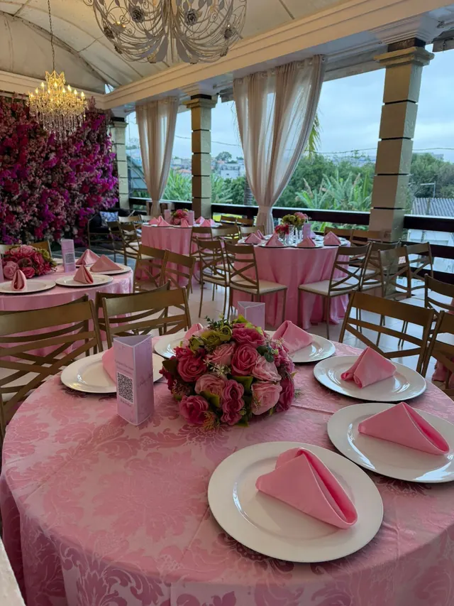 Varanda do Buffet Melvalen com mesas de toalha rosa, lustre de cristal, parede de flores e cortinas brancas
